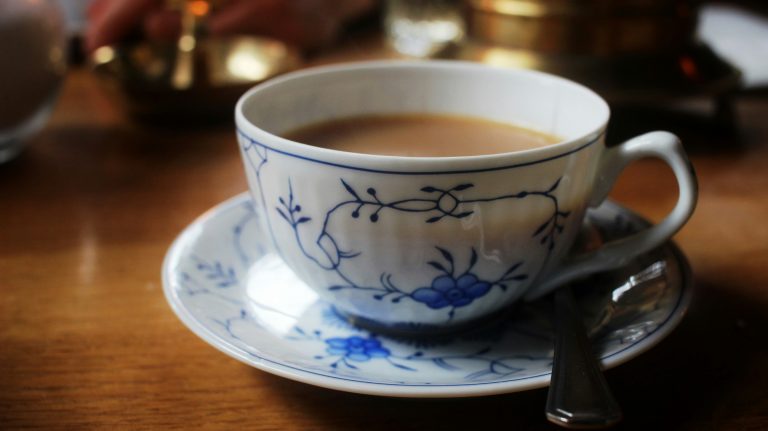 Man sieht auf dem Bild eine schöne klassische weiße ostfriesische Teetasse mit blauem Muster. Die Tasse ist mit schwarzen Tee mit Milch gefüllt.